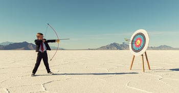 boy in business suit holding a bow and arrow and taking aim at a target on a beach.